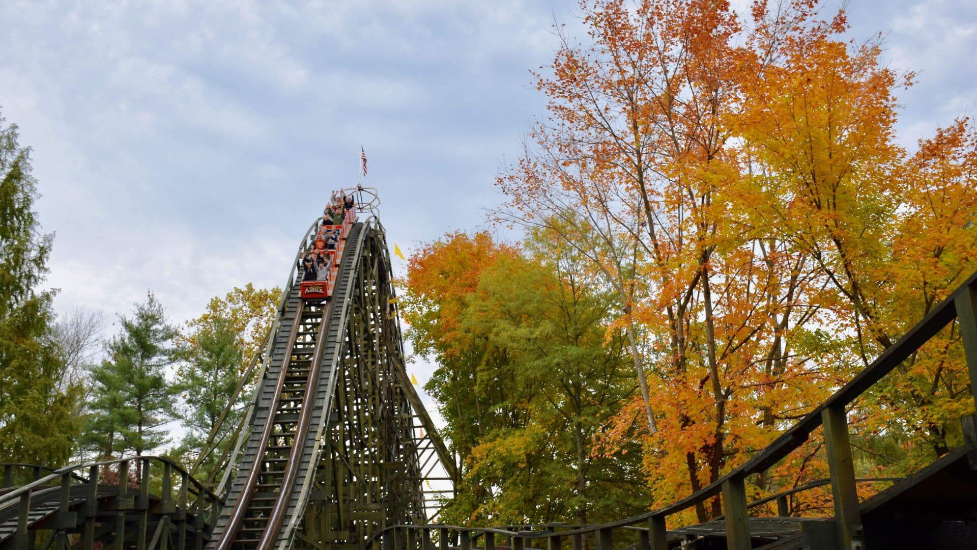 People riding cars on wooden roller coaster surrounded by orange and red trees during fall foliage season