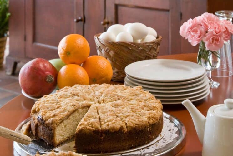 Close up view of slicked coffee cake next to a bowl of fruit, basket of eggs, small white plates, and water glasses on a round wooden table