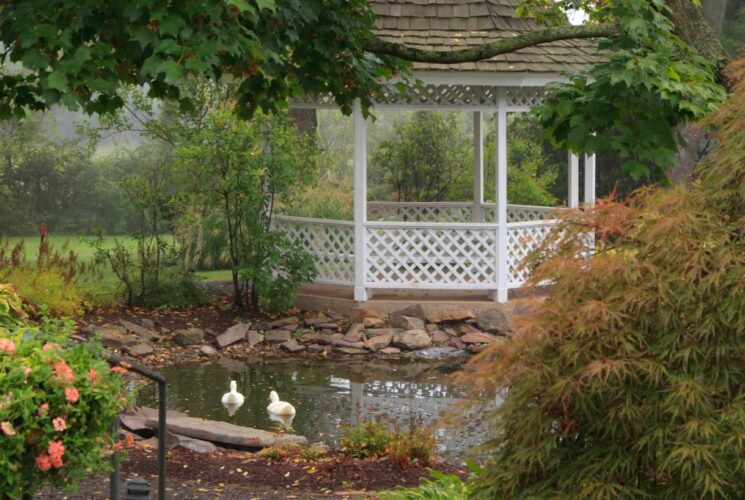 White gazebo surrounded by a small pond, green trees, green vegetation, and flowering bushes