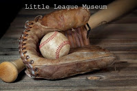 Old brown baseball mitt with dirty baseball inside, lying against wooden bat on gray wooden boards White text: Little League Museum