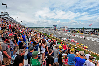 crowds of people standing in stands on left cheering yellow, red, white race cars in lead of many cars on racetrack at right