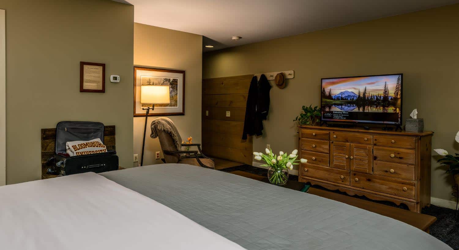 Bedroom with light taupe walls, bed with white and light sage green bedding, sitting area, and wooden dresser with large flat-screen TV