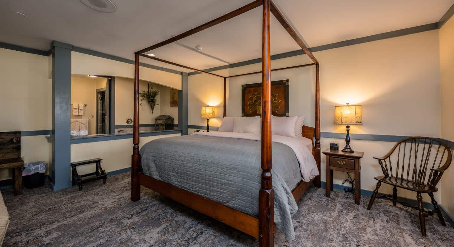 Bedroom with light cream walls, blue gray trim, multicolored carpeting, wooden canopy bed, white and light gray bedding, and sitting area