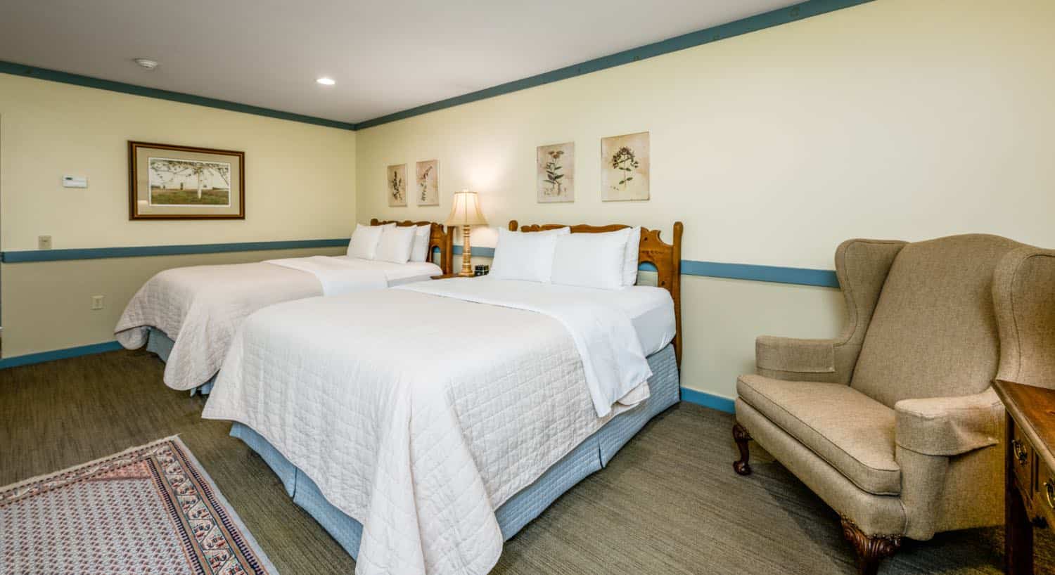 Bedroom with cream walls, blueish gray trim, two beds each with a wooden headboard and white bedding, and light tan upholstered armchair