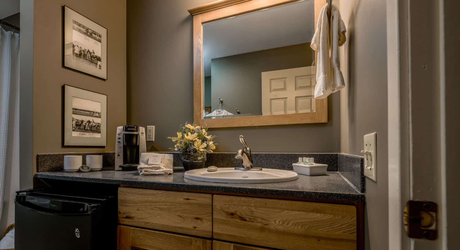 Bathroom with taupe walls, white trim, wooden vanity, quartz countertop, black mini fridge, and square wooden framed mirror