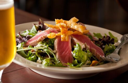 Close up view of a salad topped with grilled tuna and crispy wonton strips on a white plate next to a glass of beer