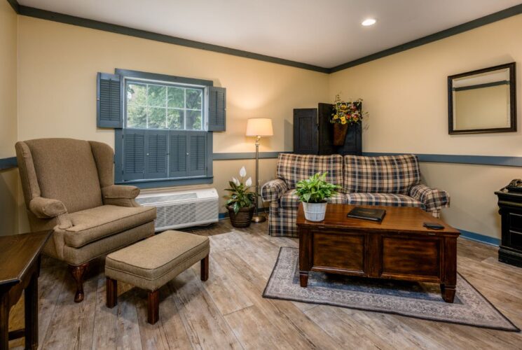 Bedroom with cream walls, blueish gray trim, hardwood flooring, tan upholstered armchair, plaid upholstered loveseat, and dark wooden coffee table