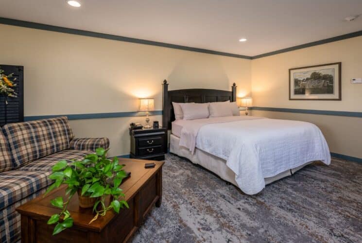 Bedroom with cream walls, blueish gray trim, dark wooden headboard, white and pale pink bedding, plaid upholstered love seat, and dark wooden coffee table