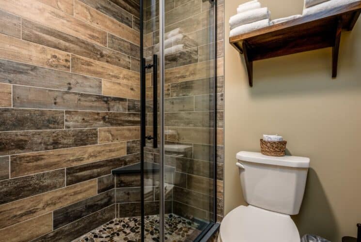 Bathroom with light tan walls, white toilet, and walk in shower with glass door, wooden tile, and pebble shower pan