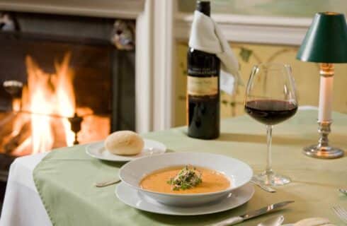 Close up of a creamy soup in a white bowl next to a white plate with a roll and class of red wine on a table with a green tablecloth and fireplace in the background