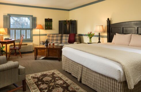 Bedroom with light yellow walls, sage green trim, light brown carpeting, dark wooden headboard, light tan and white bedding, and sitting area