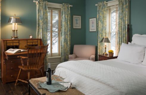 Bedroom with light blueish green walls, white trim, dark wooden headboard, white bedding, and antique wooden desk with chair