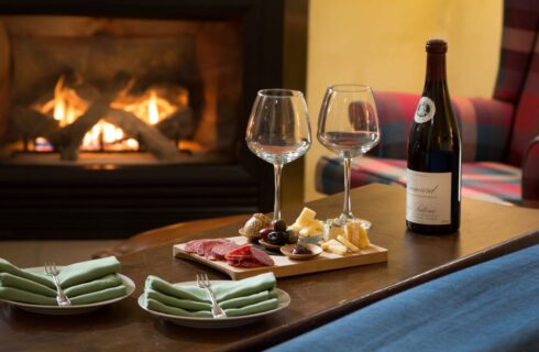 Two wine glasses on wooden cutting board with meat, cheese, and olives sitting on wooden table with fireplace in the background