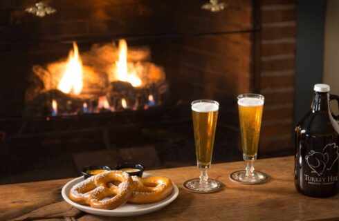 Two glasses filled with beer and white plate with homemade pretzels on wooden table with fireplace in the background