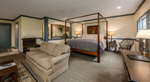 Bedroom with light yellow walls, blue gray trim, multicolored carpeting, wooden canopy bed, white and light gray bedding, and sitting area