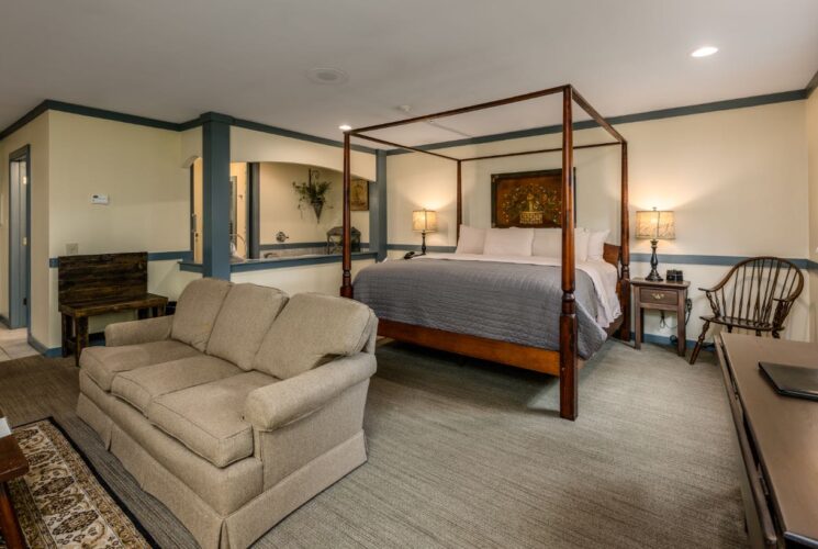 Bedroom with light yellow walls, blue gray trim, multicolored carpeting, wooden canopy bed, white and light gray bedding, and sitting area