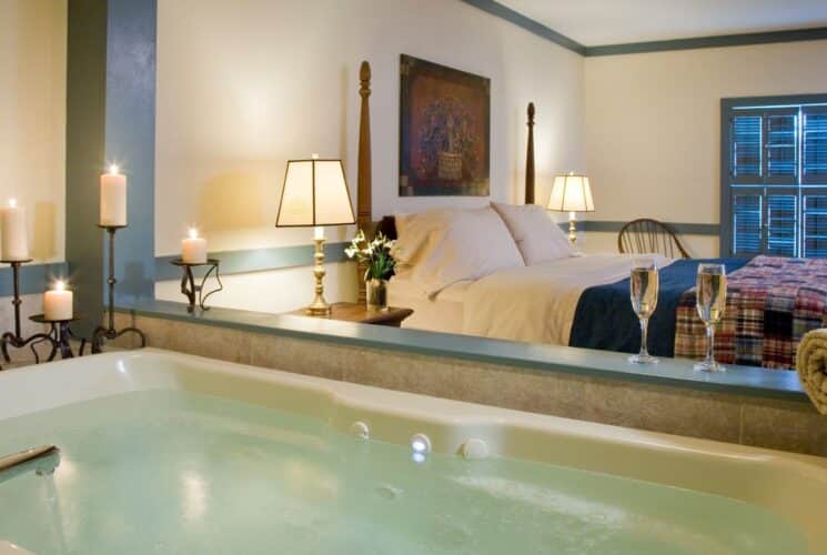 Bedroom with light yellow walls, blue gray trim, wooden four-poster bed, multicolored bedding, and large jetted tub