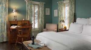 Bedroom with light blueish green walls, white trim, dark wooden headboard, white bedding, and antique wooden desk with chair