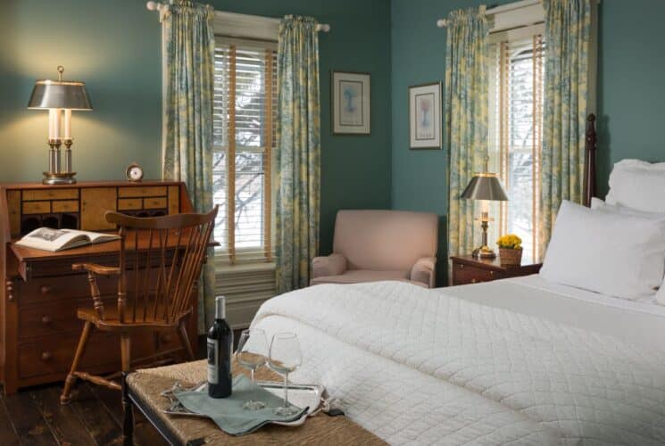 Bedroom with light blueish green walls, white trim, dark wooden headboard, white bedding, and antique wooden desk with chair