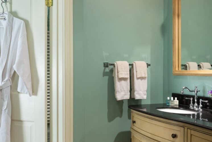 Room with light sage green walls, white trim, vanity with dark top, large wooden-framed mirror, and white robe hanging from a hook on the door