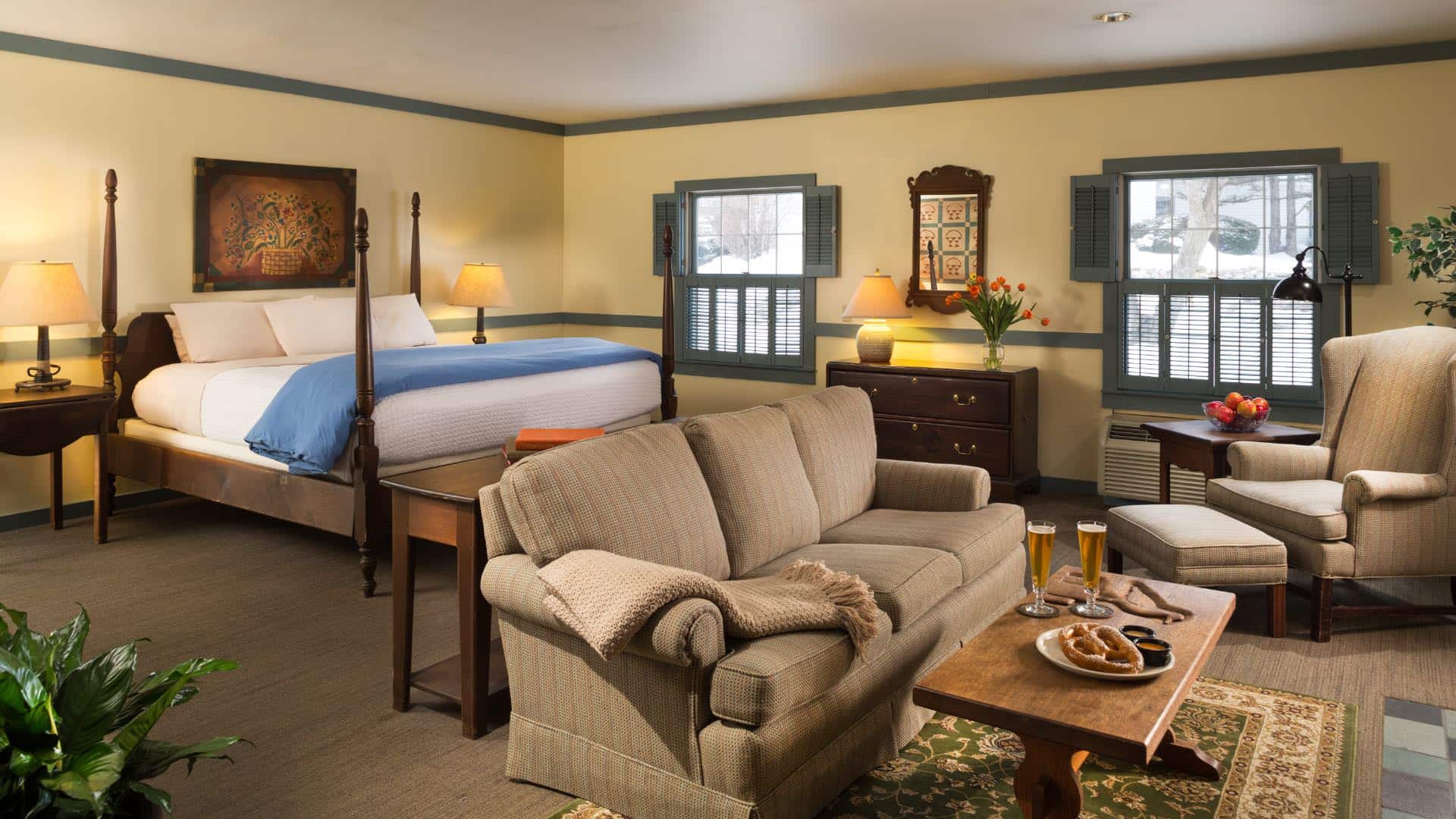 Bedroom with light yellow walls, sage green trim, dark wooden four-poster bed, white and blue bedding, and sitting area