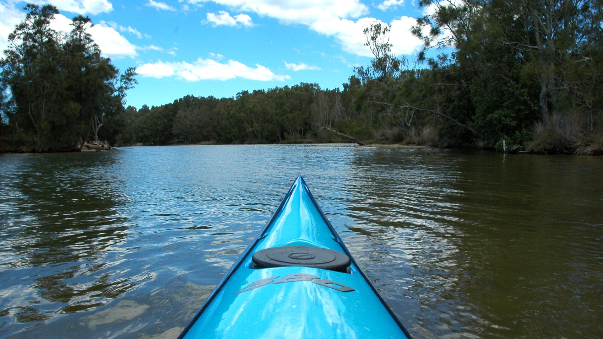 bow of turquoise kayak heading up quiet river shoreline trees & blue sky with white clouds reflected in water