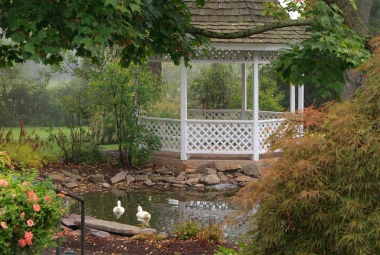 White gazebo surrounded by a small pond, green trees, green vegetation, and flowering bushes