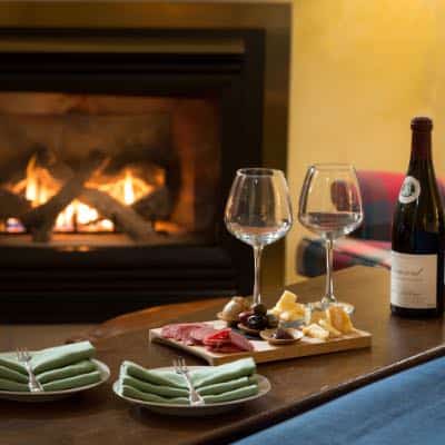 Two wine glasses on wooden cutting board with meat, cheese, and olives sitting on wooden table with fireplace in the background