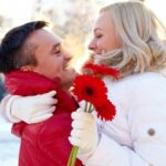 young couple cuddling on a snowy winter’s day with woman holding red gerber daisies