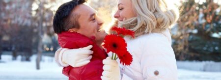 young couple cuddling on a snowy winter’s day with woman holding red gerber daisies