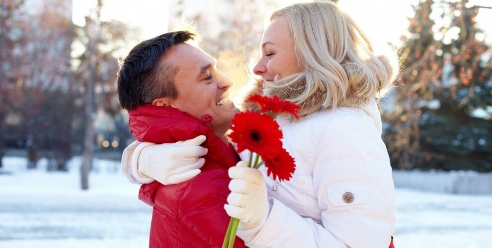 young couple cuddling on a snowy winter’s day with woman holding red gerber daisies